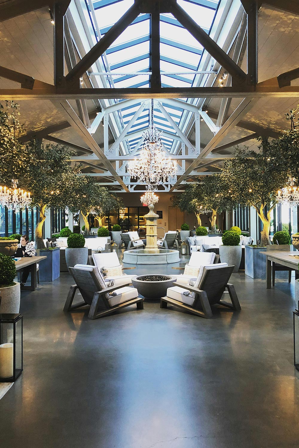 A stylish indoor lounge with skylights, chandeliers, and lush greenery.