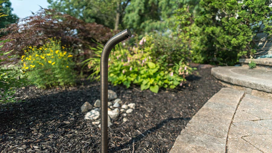 A sleek landscape lighting fixture stands among lush greenery and a stone pathway, enhancing the outdoor ambiance.