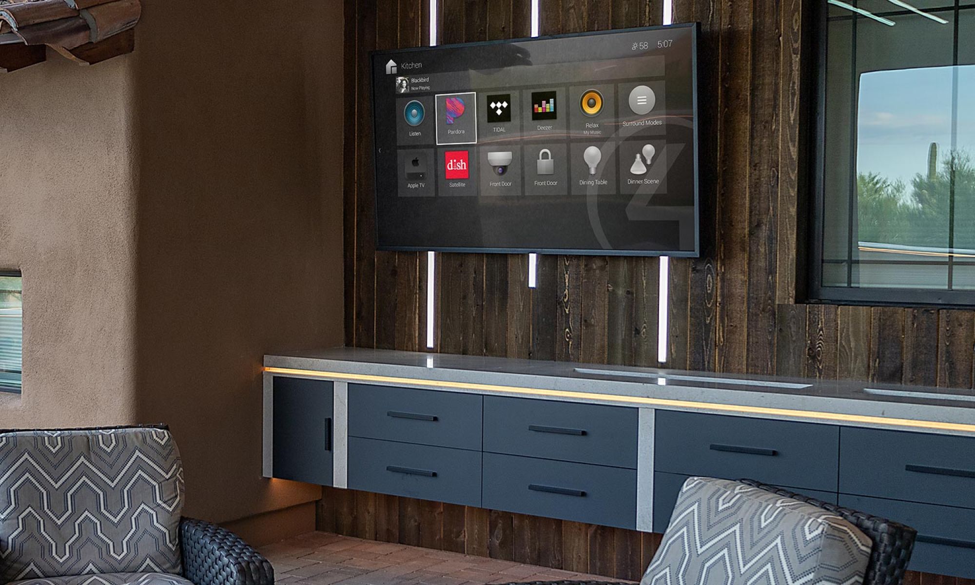 An outdoor entertainment space featuring a smart control system on a large wall-mounted screen, a built-in counter with LED lighting, and comfortable seating.
