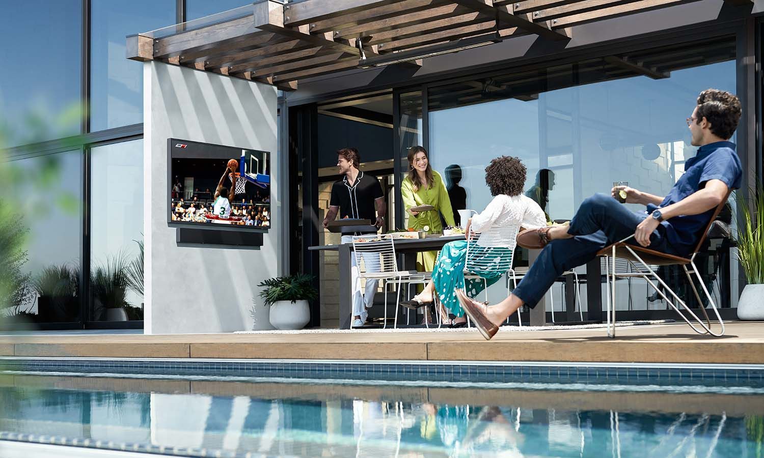 A stylish outdoor patio with a pergola, featuring a wall-mounted TV displaying a basketball game. A group of friends enjoys drinks and food by the pool, creating a lively and inviting atmosphere.