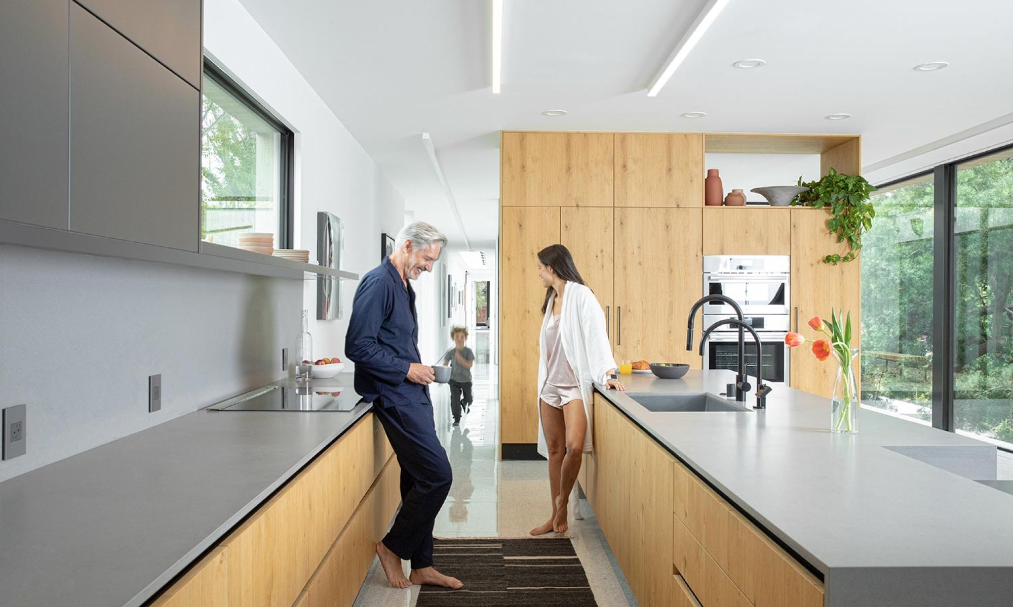 A bright, modern kitchen with natural wood cabinetry, sleek countertops, and large glass doors opening to lush greenery. A couple enjoys a casual morning while a child runs in the background.