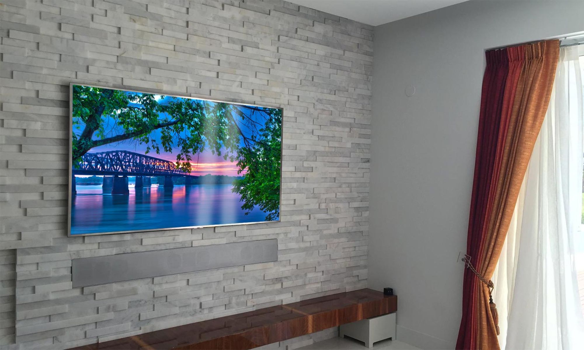 A minimalist living room with a textured gray stone wall, a mounted TV displaying a scenic sunset over a river, and a wooden bench below.