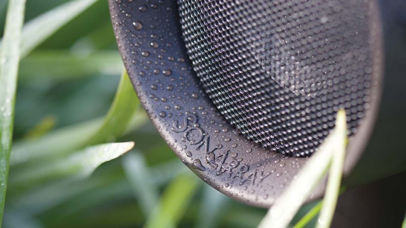 A close-up of a weather-resistant outdoor speaker covered in water droplets, surrounded by lush green grass.